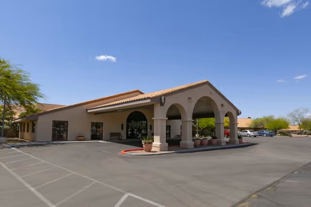 Front entrance of a single-story stucco building with an arched porte-cochère and adjacent parking lot under a clear blue sky.