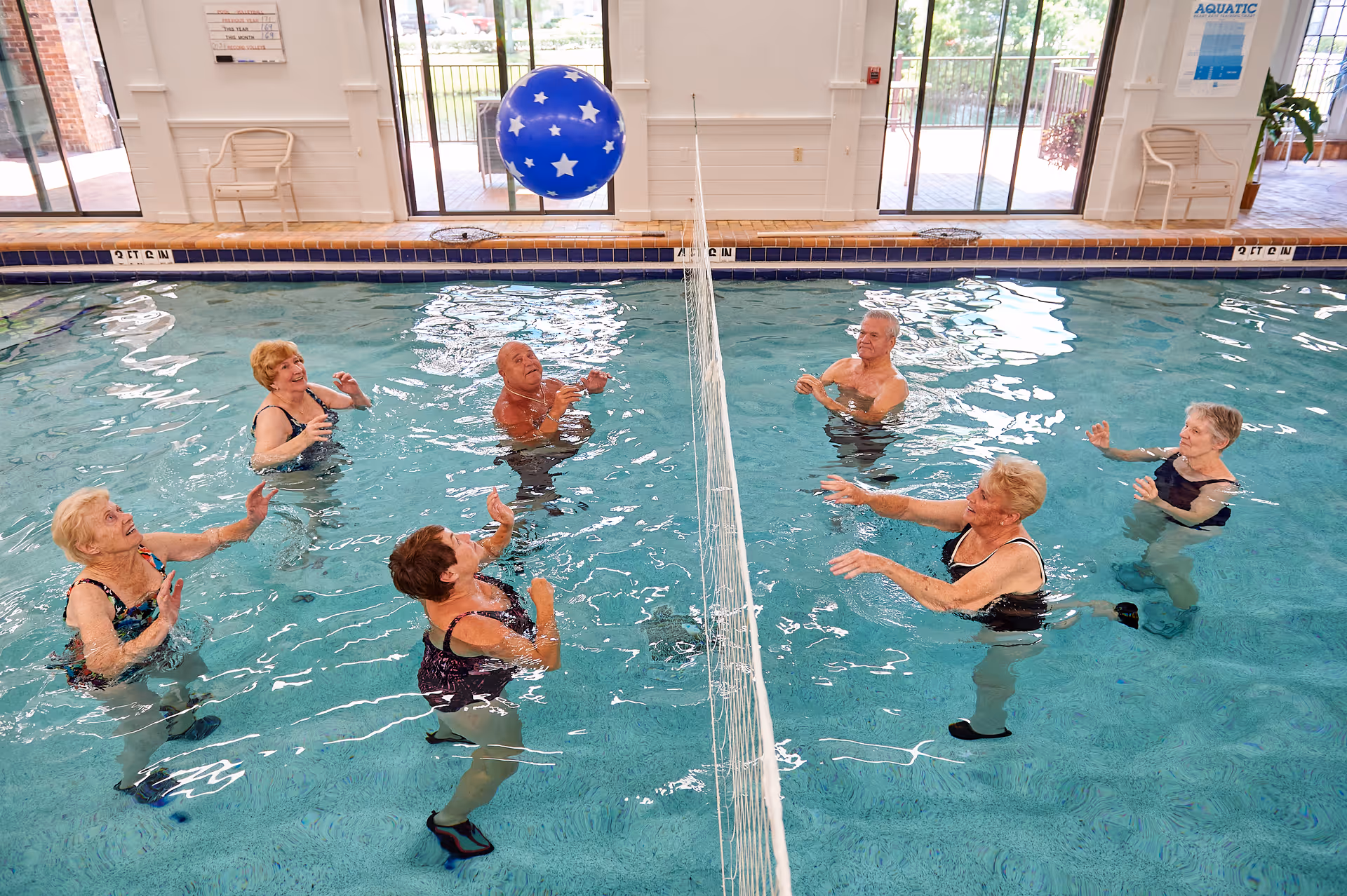 A group of older adults playing water volleyball with a large blue ball in an indoor swimming pool.