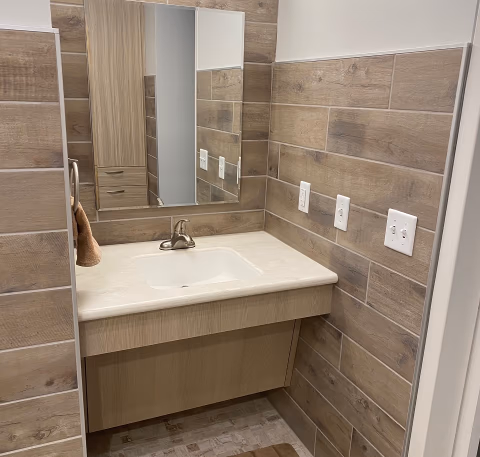 A bathroom vanity area with a rectangular sink, a single-handle faucet, and a large mirror above. The walls are covered with wood-look tiles, and there are multiple electrical outlets and light switches on the right wall. A brown towel hangs on a ring on the left side.