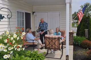 Four elderly people sitting and chatting on a covered patio outside a residential building with white siding. There are flowers and greenery around the patio, and an American flag is visible in the background.