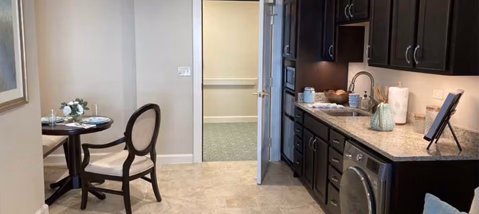 Interior view of a kitchen and small dining area in a senior living facility. The kitchen features dark wood cabinets, a granite countertop, a sink, and built-in appliances including a microwave and a washing machine. To the left, there is a small round dining table set for two with plates, glasses, and a floral centerpiece. An open door leads to a hallway with carpeted flooring.