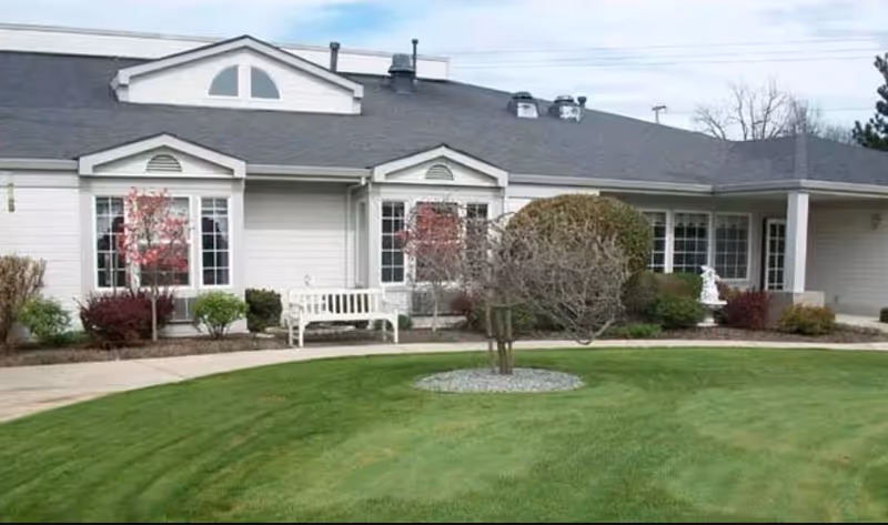 Exterior view of a single-story building with white siding and a dark gray roof, featuring multiple windows and a small landscaped garden with trimmed bushes, a tree, and a white bench in front.
