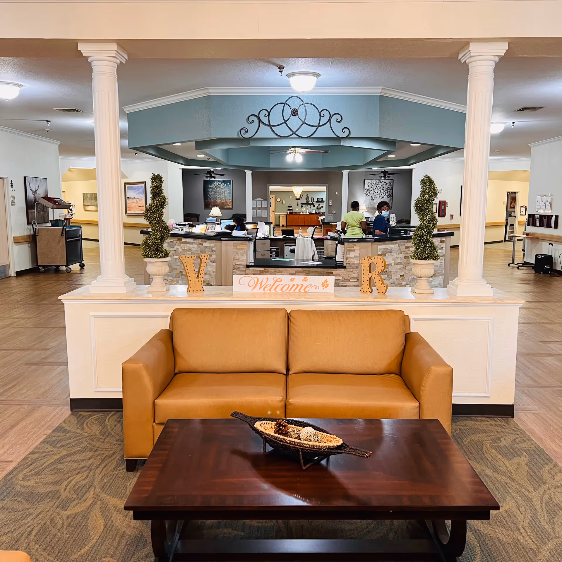 Interior view of a nursing and rehabilitation center reception area with a tan couch and dark wooden coffee table in the foreground. Behind the couch is a low wall with decorative letters 'V' and 'R' and a sign that says 'Welcome'. The reception desk is staffed by two people wearing masks, with decorative plants and artwork on the walls. The ceiling has a blue octagonal design with a ceiling fan and lighting.
