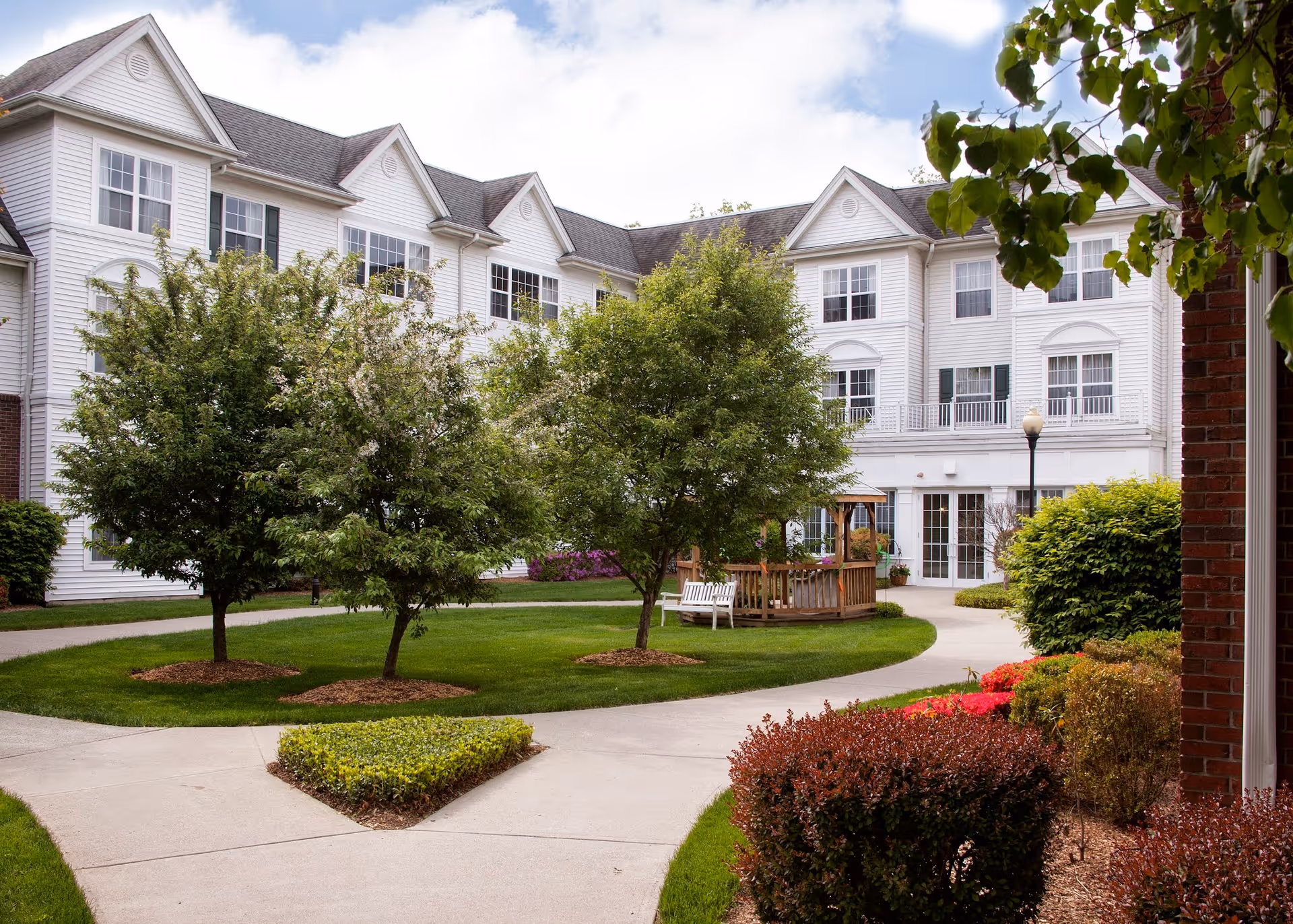 Well-maintained courtyard with trees, walkways, a gazebo and benches in front of a white multi-story senior living building.