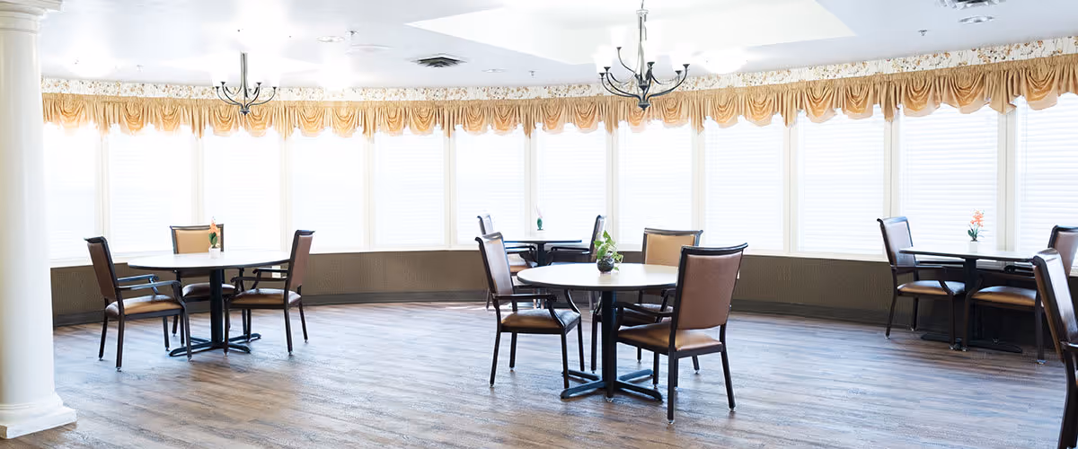 A bright dining room with multiple round tables and chairs arranged neatly. The room features large windows with white blinds and a decorative valance, wooden flooring, and two ceiling chandeliers.