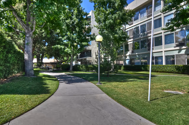 A paved walkway curves through a grassy outdoor area with trees and lamp posts, adjacent to a multi-story building with large windows under a clear blue sky.