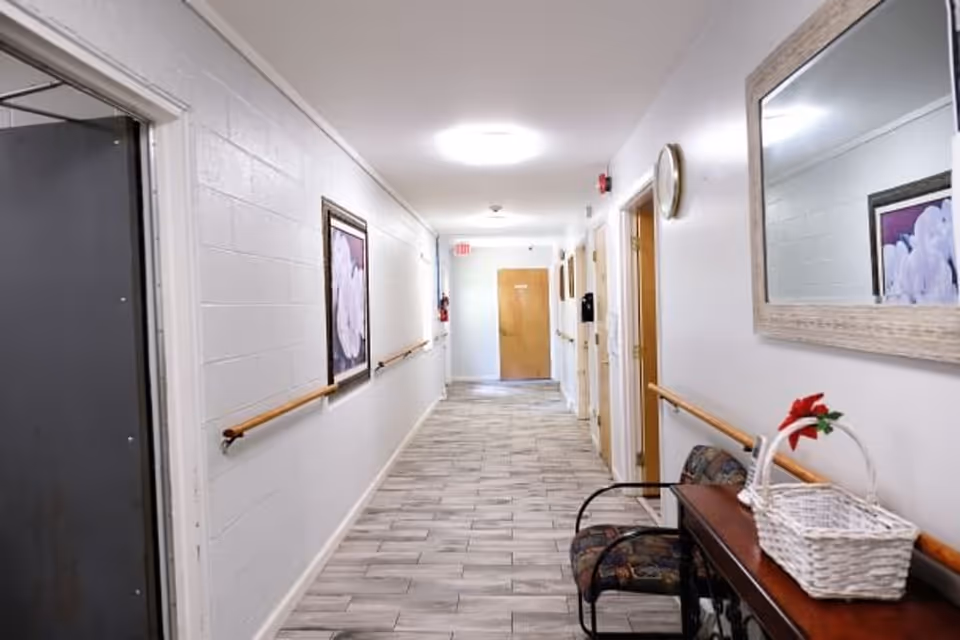 A well-lit hallway in a senior living facility with light-colored walls and tiled flooring. The hallway features wooden handrails on both sides, a framed floral picture on the left wall, a large mirror on the right wall, a small table with a white wicker basket and a chair underneath it. Several doors line the right side of the hallway, and an exit sign is visible at the far end.
