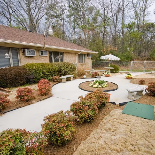 Outdoor courtyard area at a senior living facility with a concrete walkway surrounded by bushes and flower beds. There are stone benches, a round flower bed in the center, a table with an umbrella, and a chain-link fence enclosing the area. The building exterior is visible with windows and air conditioning units.