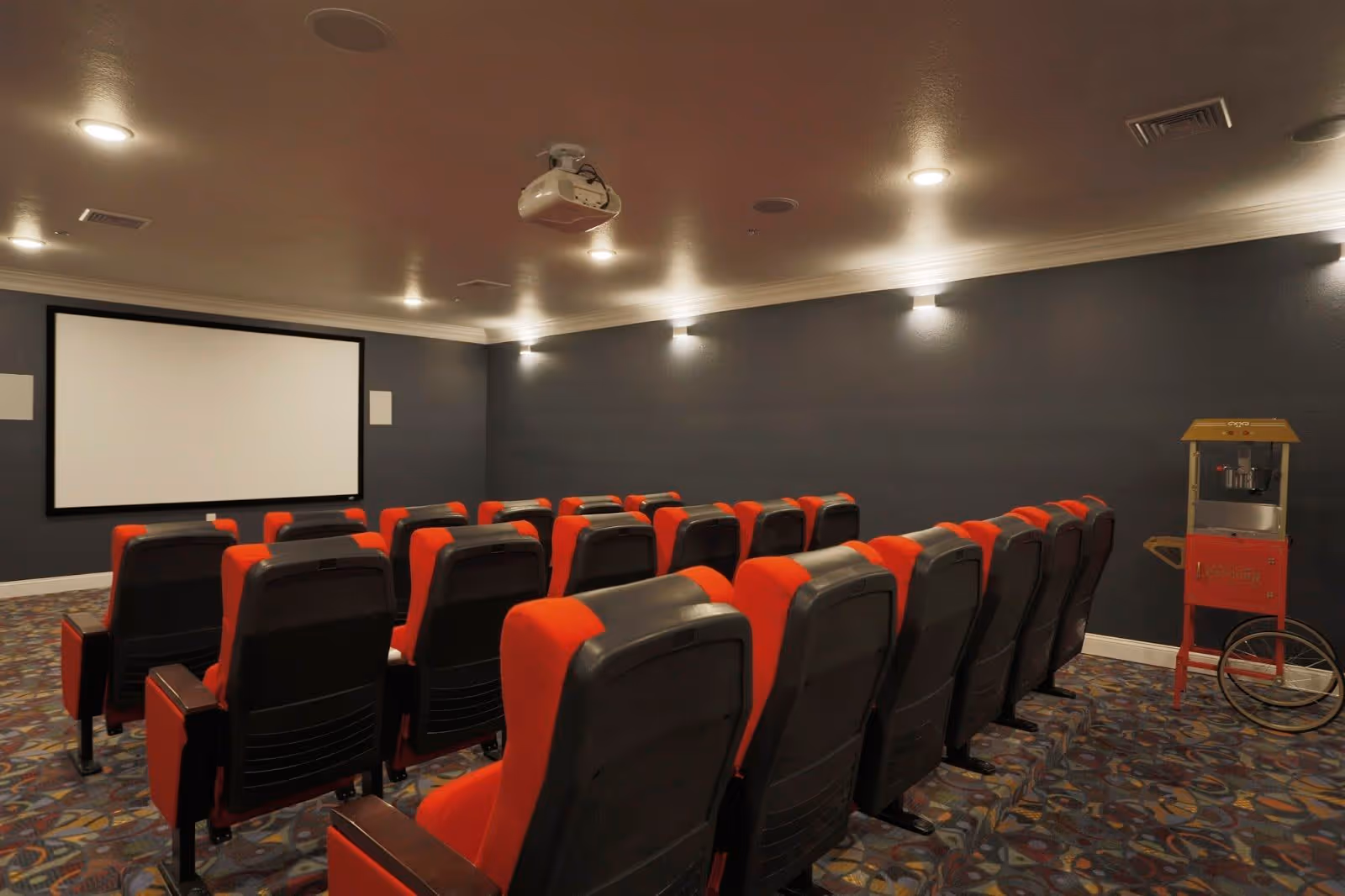 A small theater room with rows of red and black cushioned seats facing a large blank projection screen on a dark gray wall. The room has a patterned carpet floor, ceiling lights, a ceiling-mounted projector, and a vintage-style popcorn machine on the right side.