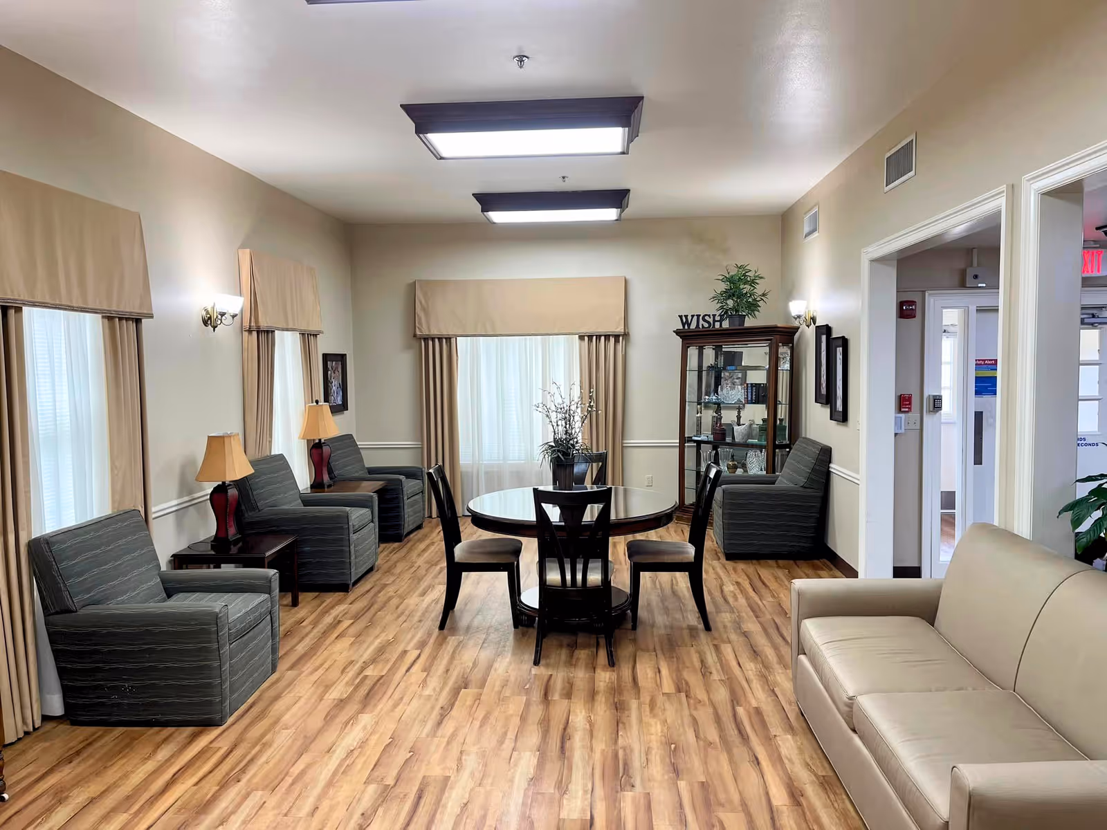 A well-lit common area with wood flooring featuring a round table with four chairs in the center, several armchairs along the walls, a beige sofa on the right, and a glass display cabinet with decorative items and a plant on top. The room has beige walls, windows with beige curtains, and ceiling lights.