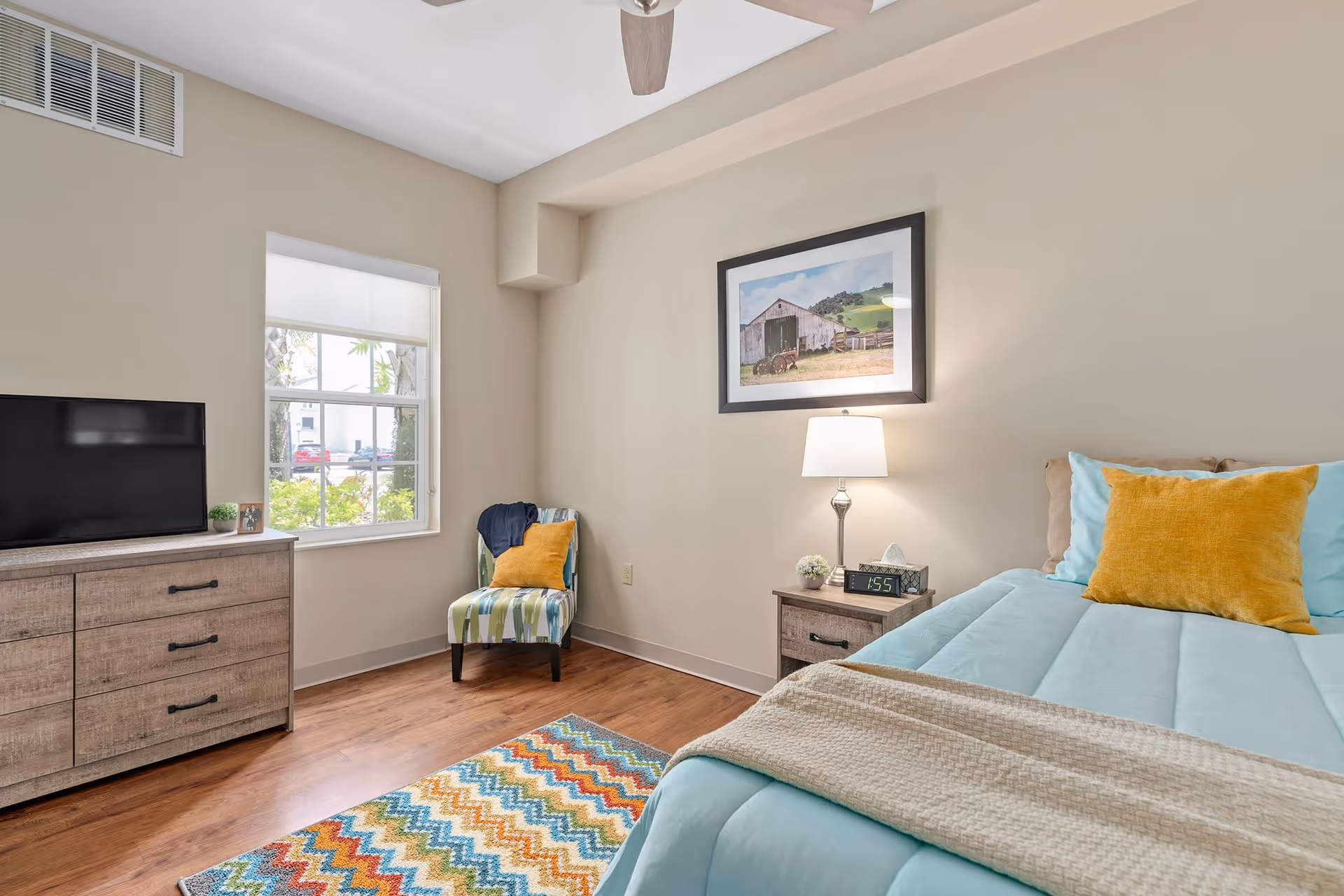 A cozy bedroom with a bed featuring light blue bedding and a mustard yellow pillow. Next to the bed is a wooden nightstand with a lamp, a digital clock, and a small plant. A framed picture of a barn hangs on the beige wall above the nightstand. Across from the bed is a wooden dresser with a flat-screen TV on top. A window with a white blind lets in natural light, and a colorful zigzag patterned rug lies on the wooden floor. A striped chair with a mustard yellow pillow is placed near the window.