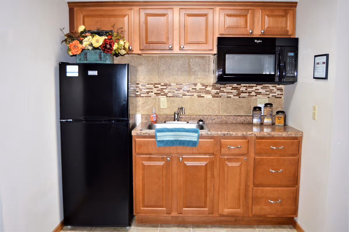 A compact kitchen area featuring wooden cabinets, a black refrigerator with a floral arrangement on top, a countertop with a sink and a blue towel hanging over the sink, a black microwave mounted above the counter, and three decorative canisters on the right side of the counter.