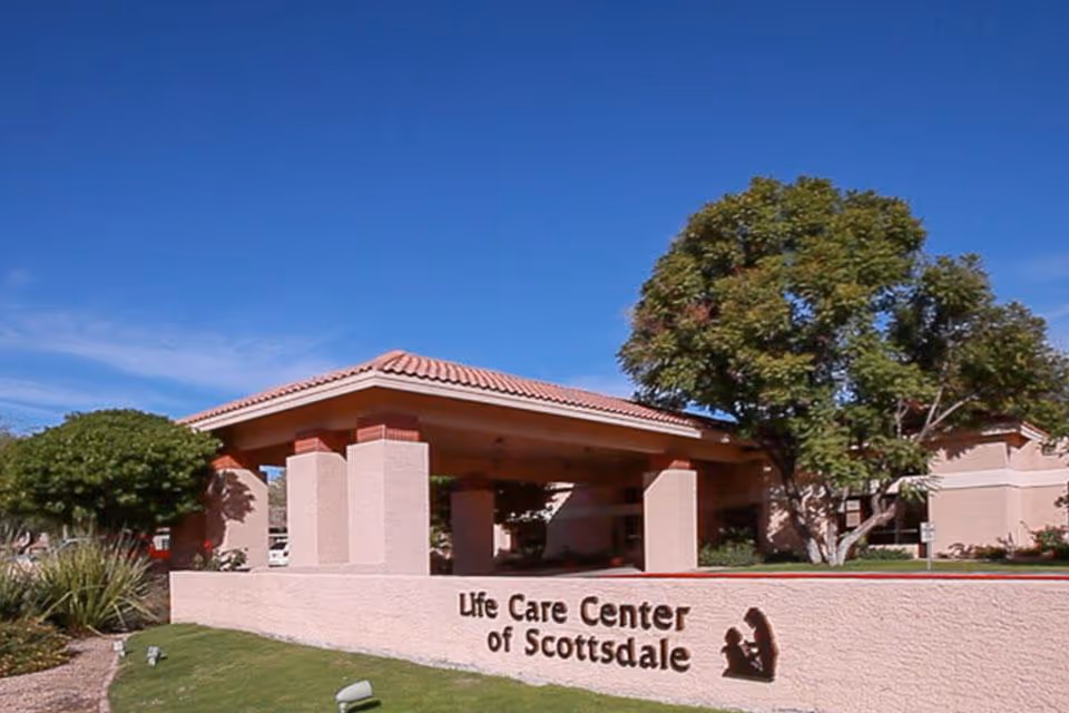Exterior view of Life Care Center of Scottsdale building with a covered entrance, surrounded by trees and landscaping under a clear blue sky.
