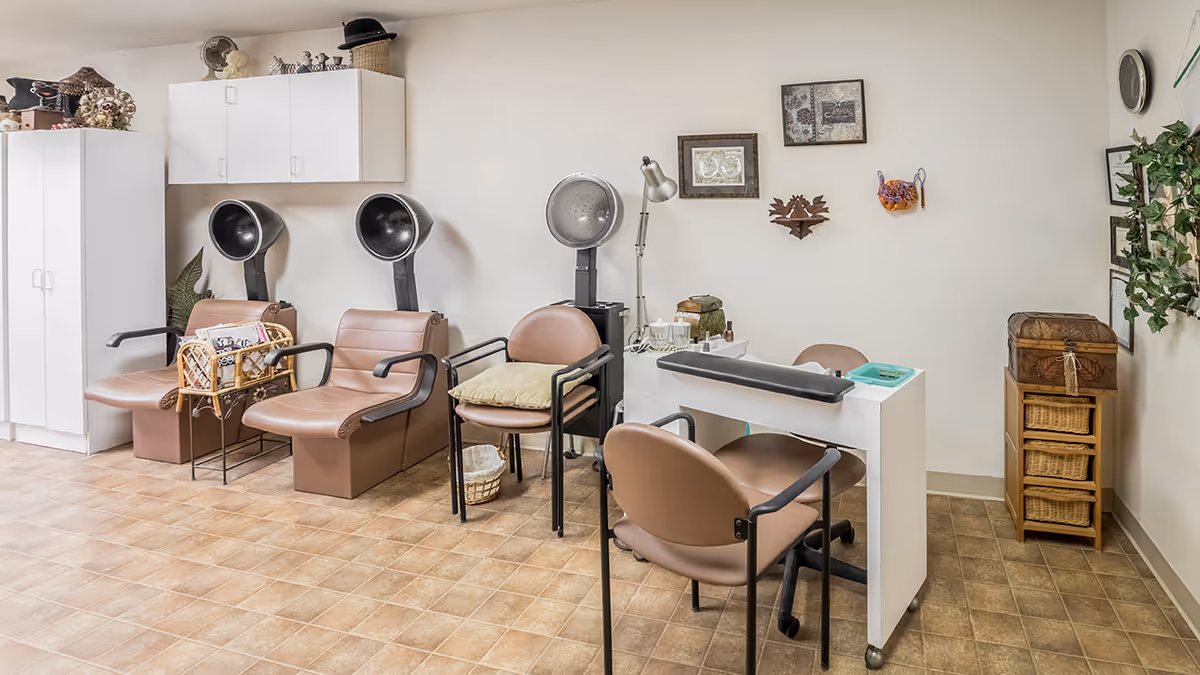 Interior view of a salon area in a senior living facility featuring two brown salon chairs with hair dryers, a small table with a lamp and manicure supplies, and several brown chairs. The walls are decorated with framed pictures and a clock, and there are storage cabinets and a small shelving unit with baskets.