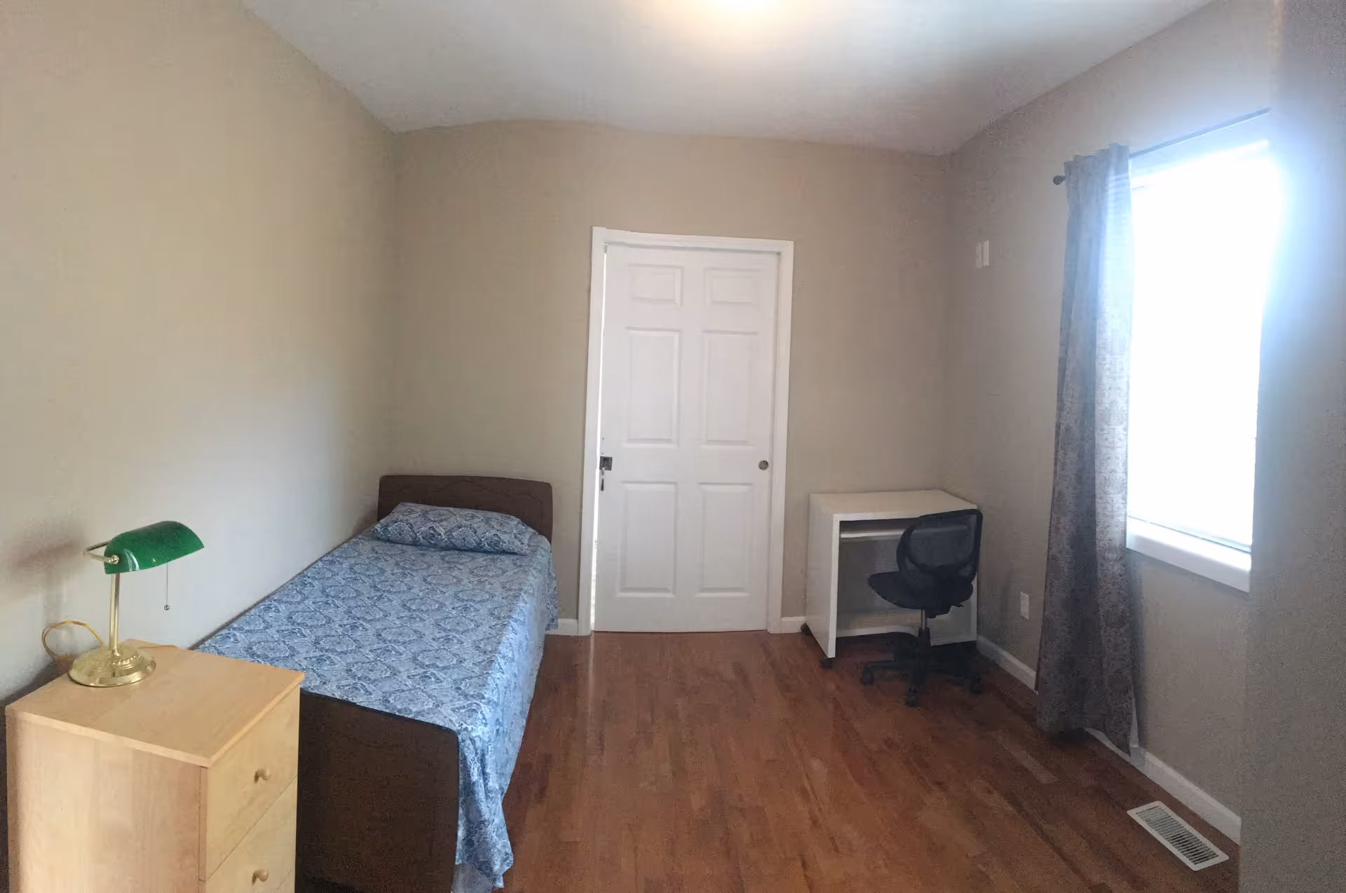 A small bedroom with a single bed covered in a blue patterned bedspread, a wooden nightstand with a green desk lamp, a white desk with a black office chair, a window with patterned curtains, and a closed white door. The room has beige walls and wooden flooring.