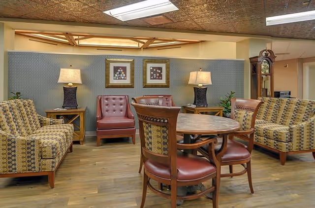 A cozy common area with patterned sofas, two red leather armchairs, a round wooden table with four chairs, two table lamps on side tables, two framed floral artworks on the wall, and a tall grandfather clock in the corner.