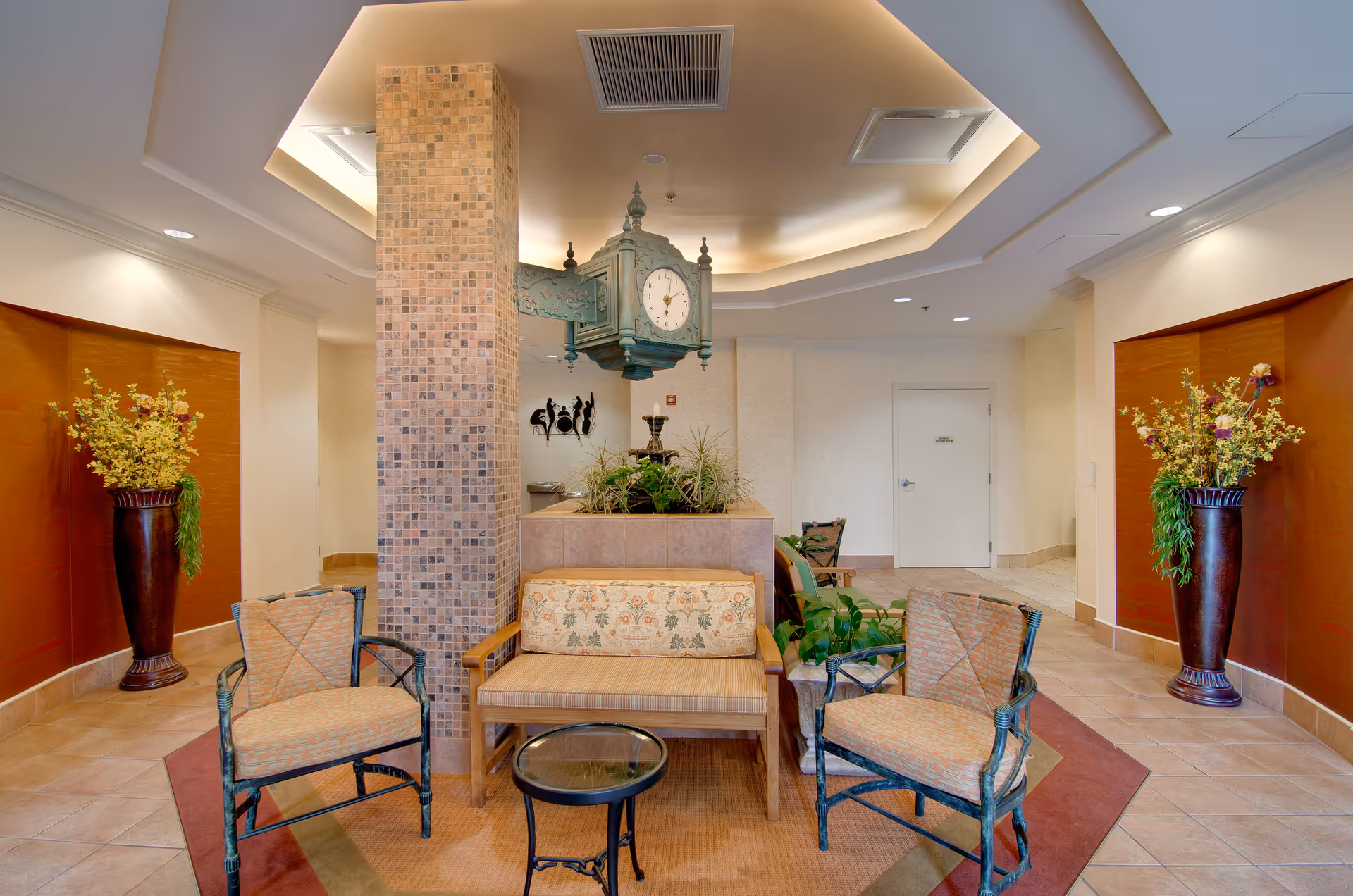 A cozy seating area in a senior living facility lobby featuring two cushioned armchairs and a cushioned bench around a small round glass-top table. The space has a tiled floor, a mosaic-tiled column, and a decorative hanging clock above a planter with greenery. Large vases with yellow and purple flowers flank the seating area against warm-toned wall panels.