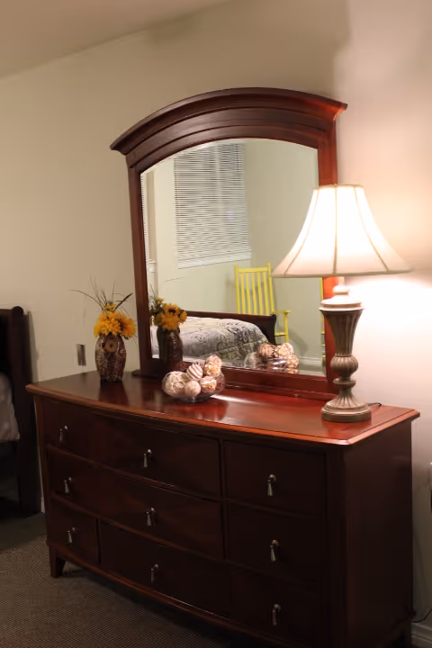 A wooden dresser with nine drawers and a large matching mirror. On top of the dresser are two vases with yellow flowers, a decorative bowl with seashells, and a lit table lamp. The mirror reflects part of a bed, a window with blinds, and a yellow chair.