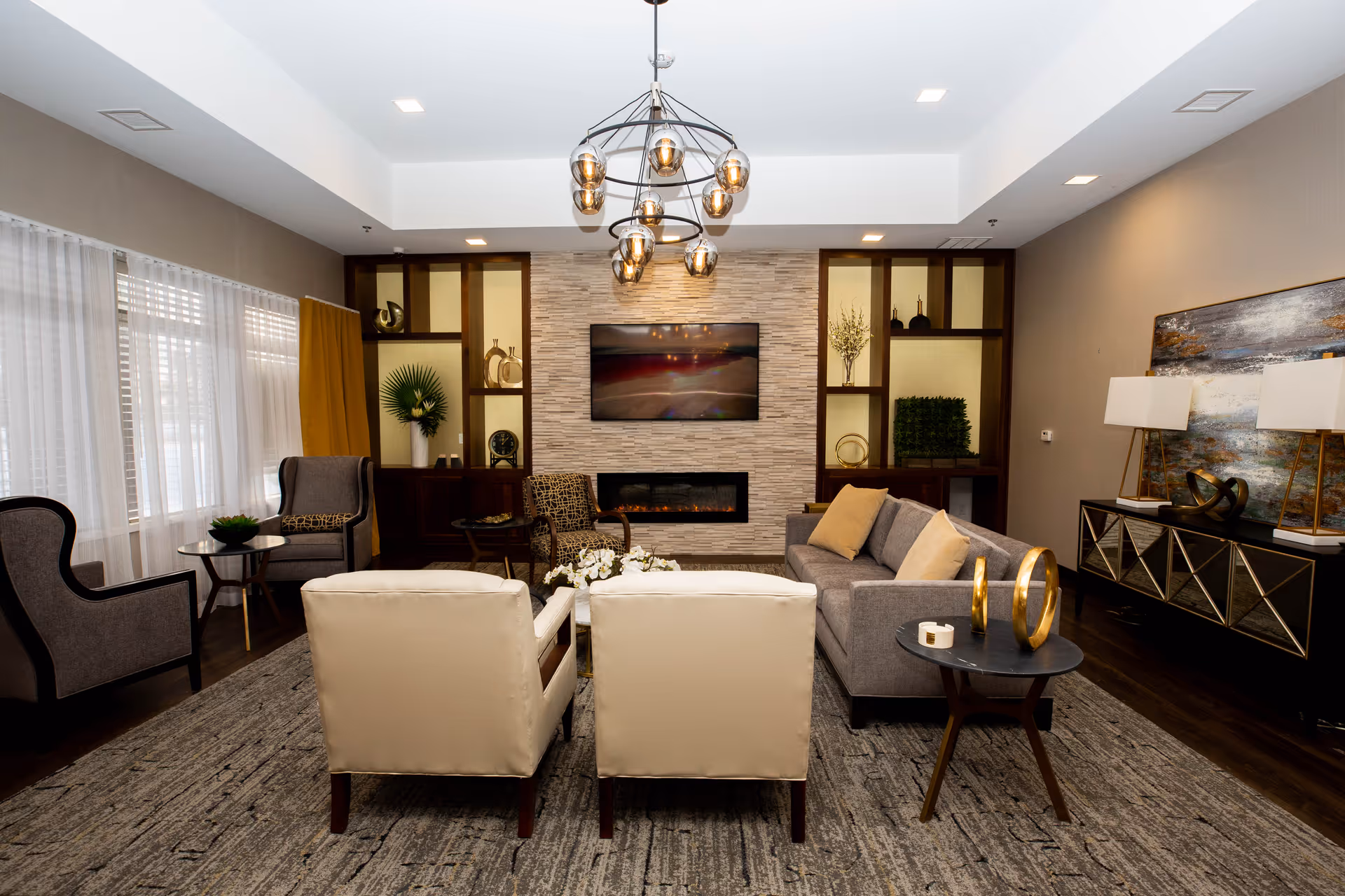 A modern living room with a gray sofa, two beige armchairs, and two dark gray armchairs arranged around a coffee table on a large area rug. The room features a stone accent wall with a mounted flat-screen TV above a built-in electric fireplace. Built-in shelves with decorative items flank the fireplace. A contemporary chandelier hangs from the ceiling, and a sideboard with two lamps and artwork is visible on the right side of the room. Large windows with sheer white curtains and mustard yellow drapes are on the left.