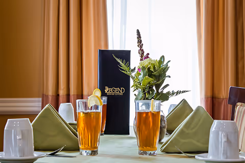 A dining table set with two glasses of iced tea, folded napkins, cups, a floral centerpiece and a standing menu in front of a window.