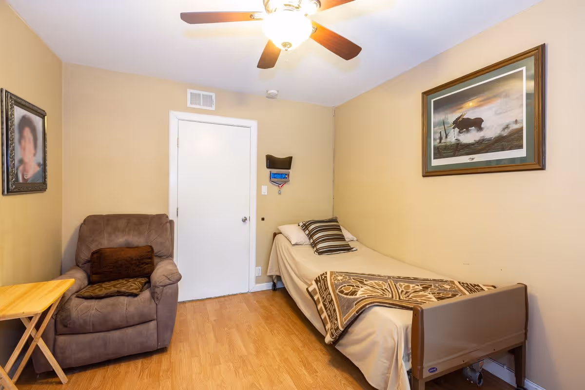 Small bedroom with a single bed along the right wall, a recliner and folding side table on the left, wood flooring and a ceiling fan.