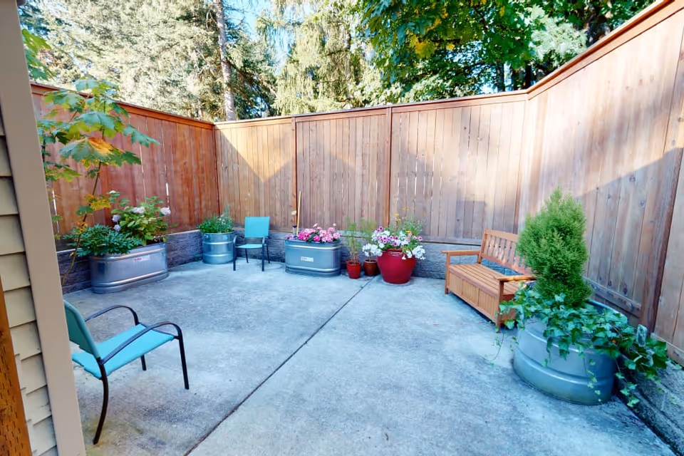 Small enclosed outdoor patio with a wooden fence, concrete floor, potted plants, chairs, and a wooden bench.