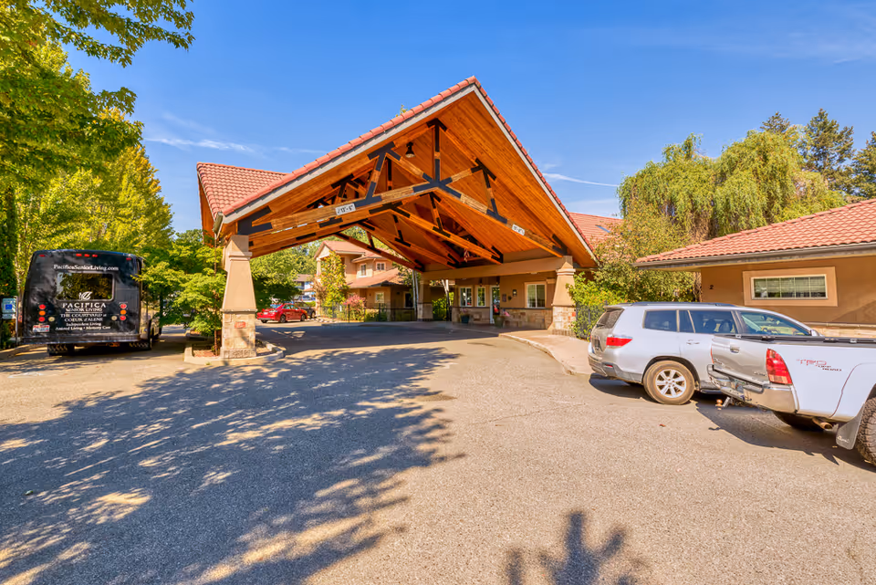 Covered entrance/porte-cochere of a senior living facility with a wooden peaked canopy, parked cars, and a shuttle bus.