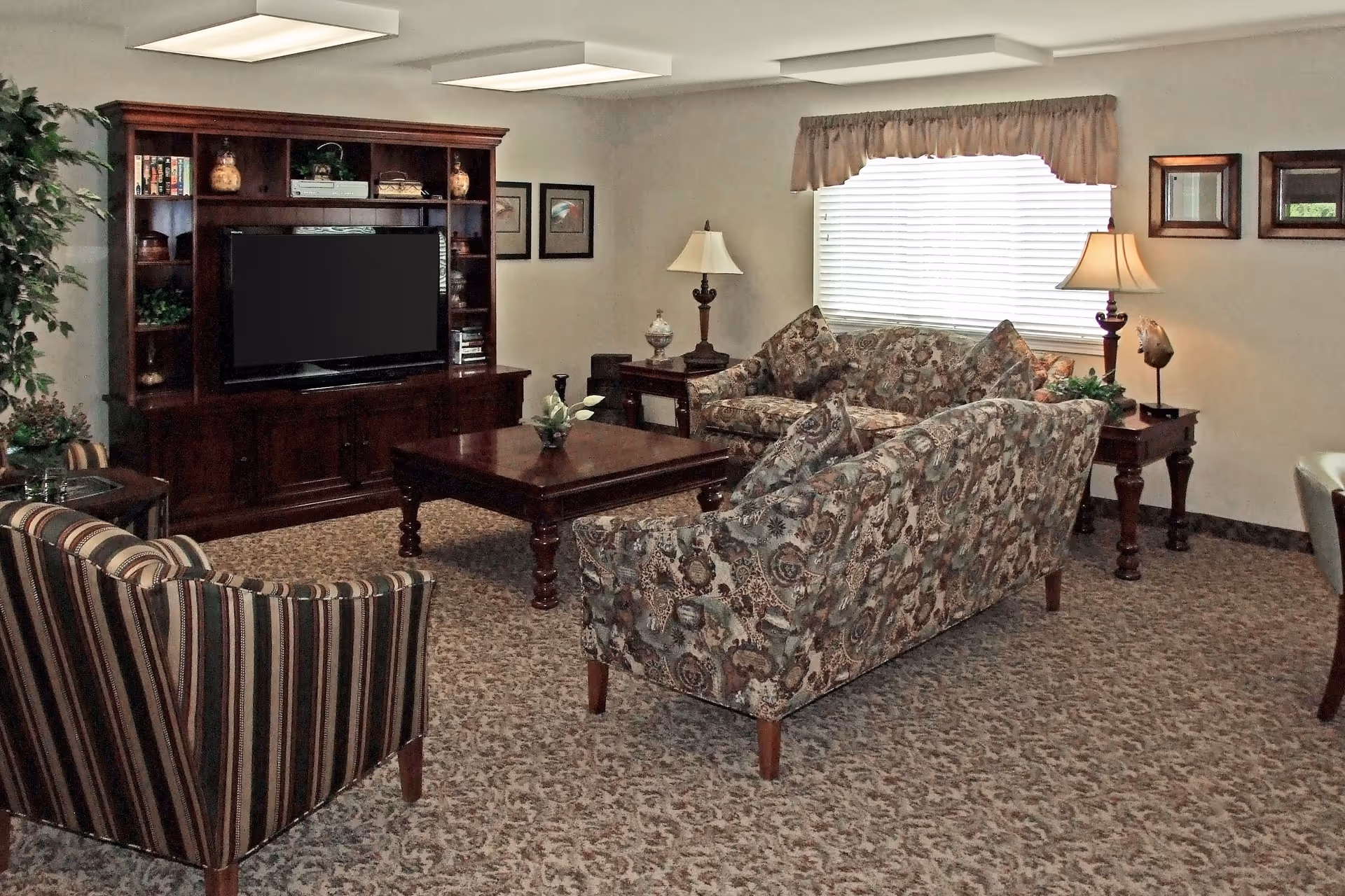 A cozy living room with patterned sofas and a striped armchair arranged around a wooden coffee table. A large wooden entertainment center with a flat-screen TV and decorative items is against the wall. Two table lamps are on side tables near a window with blinds and a valance. The room has carpeted flooring and framed pictures on the walls.
