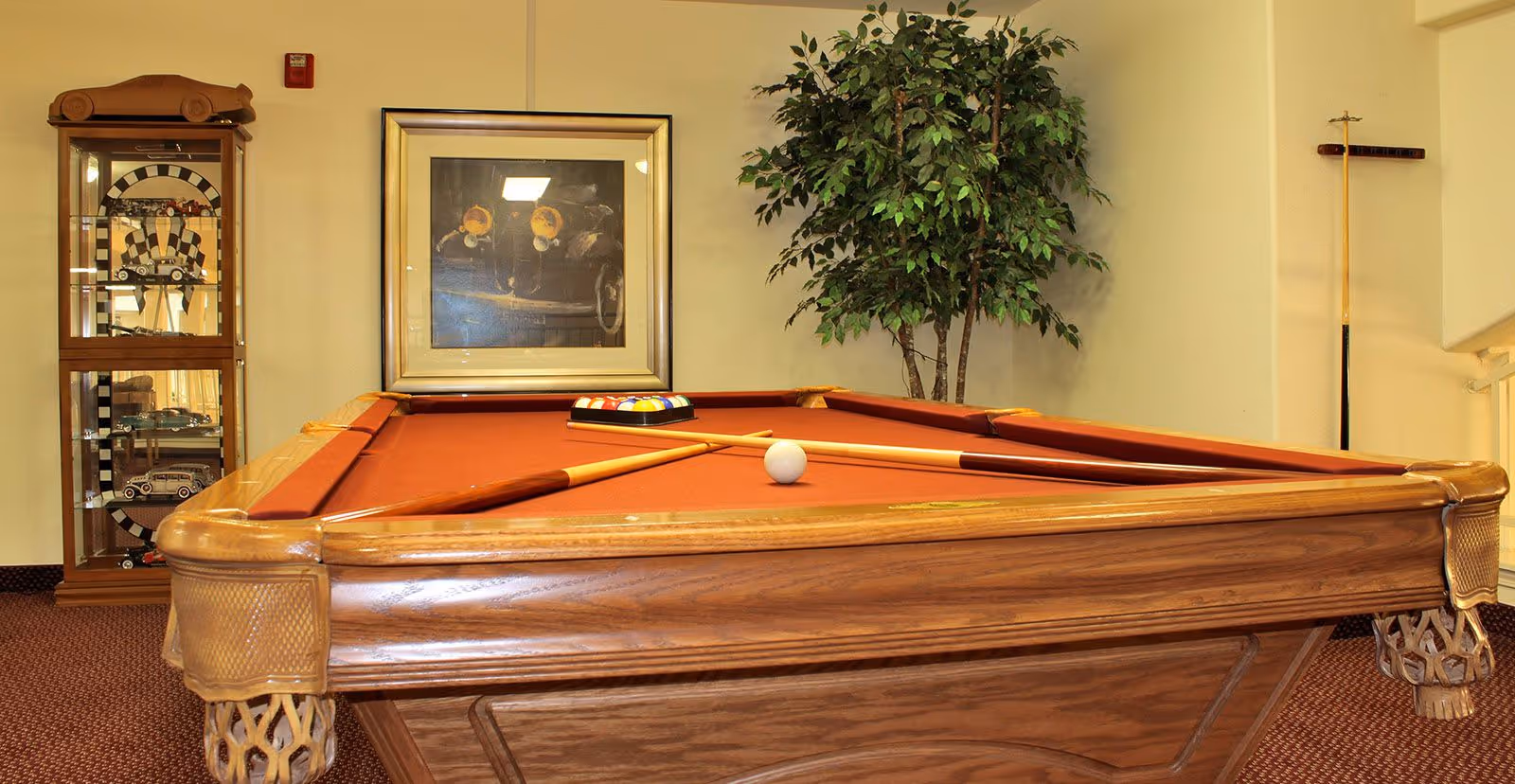 A pool table with a red felt surface, two pool cues crossed on top, and a rack of billiard balls placed near the far end. Behind the pool table is a framed picture of a vintage car, a tall green potted plant, and a wooden display cabinet with model cars inside. The room has beige walls and carpeted flooring.