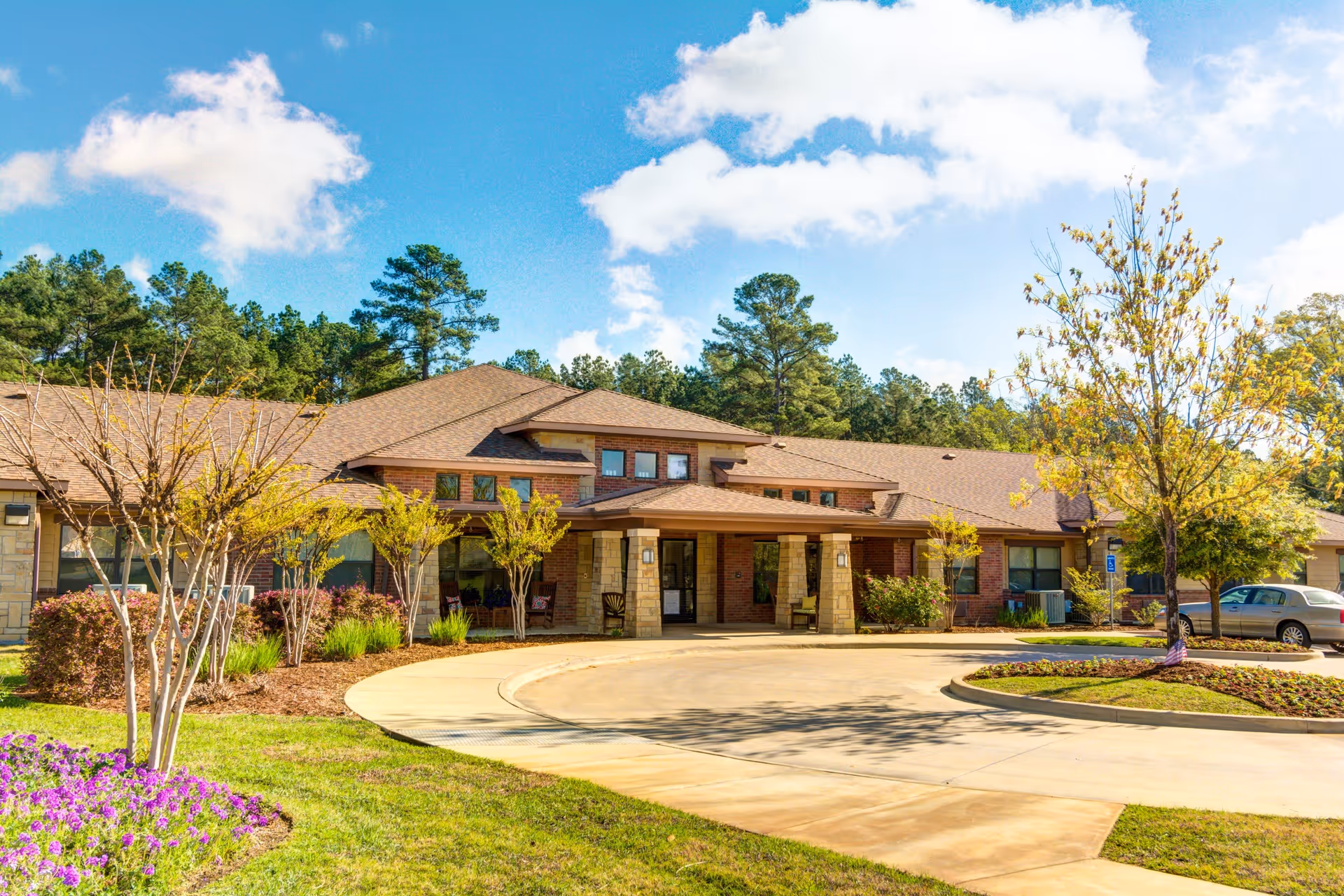Exterior view of Oak Hills Terrace Memory Care facility showing a single-story building with a covered entrance, surrounded by landscaped greenery, trees, and a circular driveway under a blue sky with scattered clouds.