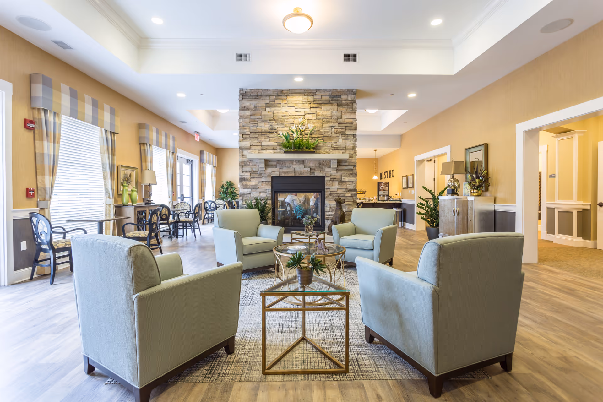 A bright and spacious living room area in an assisted living facility featuring four light green armchairs arranged around a glass coffee table on a patterned rug. A stone fireplace with a plant on the mantel is centered in the background. Large windows with striped valances allow natural light to fill the room. Additional seating with small tables lines the left wall, and a hallway and buffet area are visible on the right.