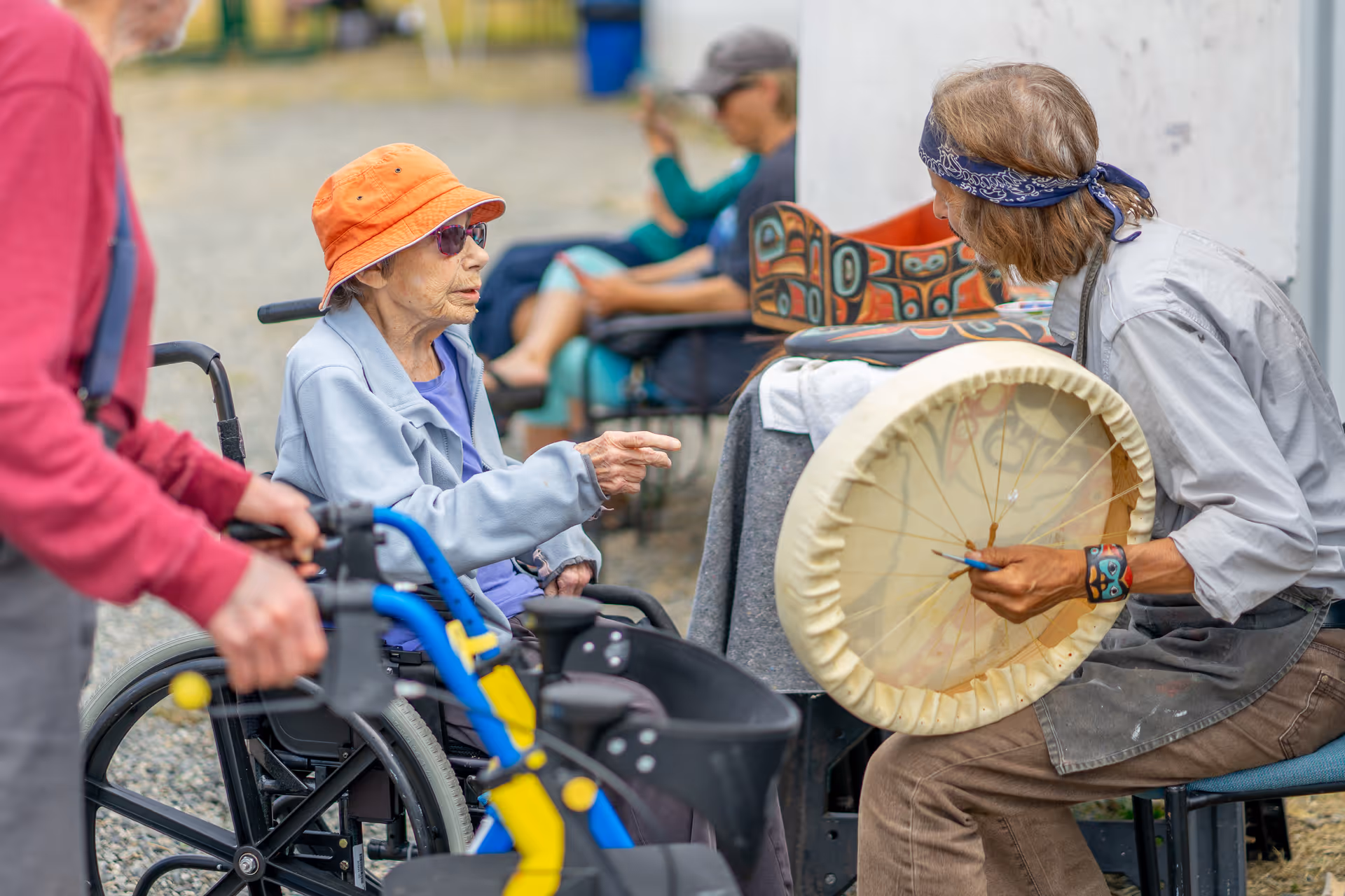 An elderly woman in a wheelchair wearing an orange hat and sunglasses is interacting with a person playing a large hand drum outdoors. Other people are seated in the background, and the setting appears to be a casual outdoor gathering.