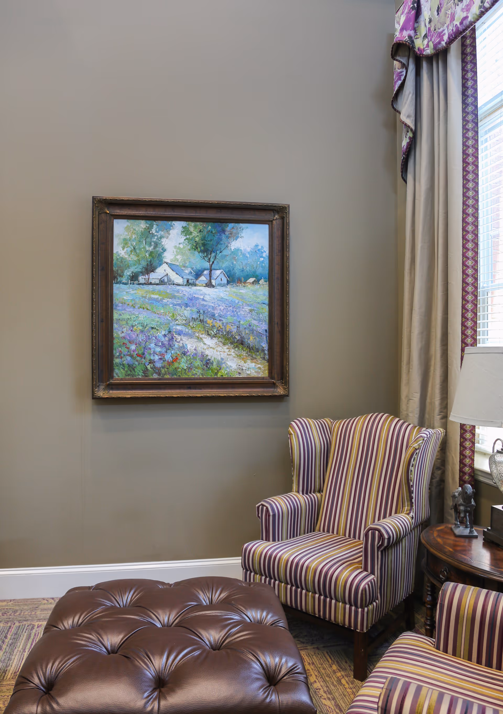 A cozy corner of a living room featuring a striped upholstered armchair and matching ottoman, a wooden side table with a lamp and small statue, a window with beige curtains and purple valance, and a framed painting of a countryside scene with trees and houses on the wall.