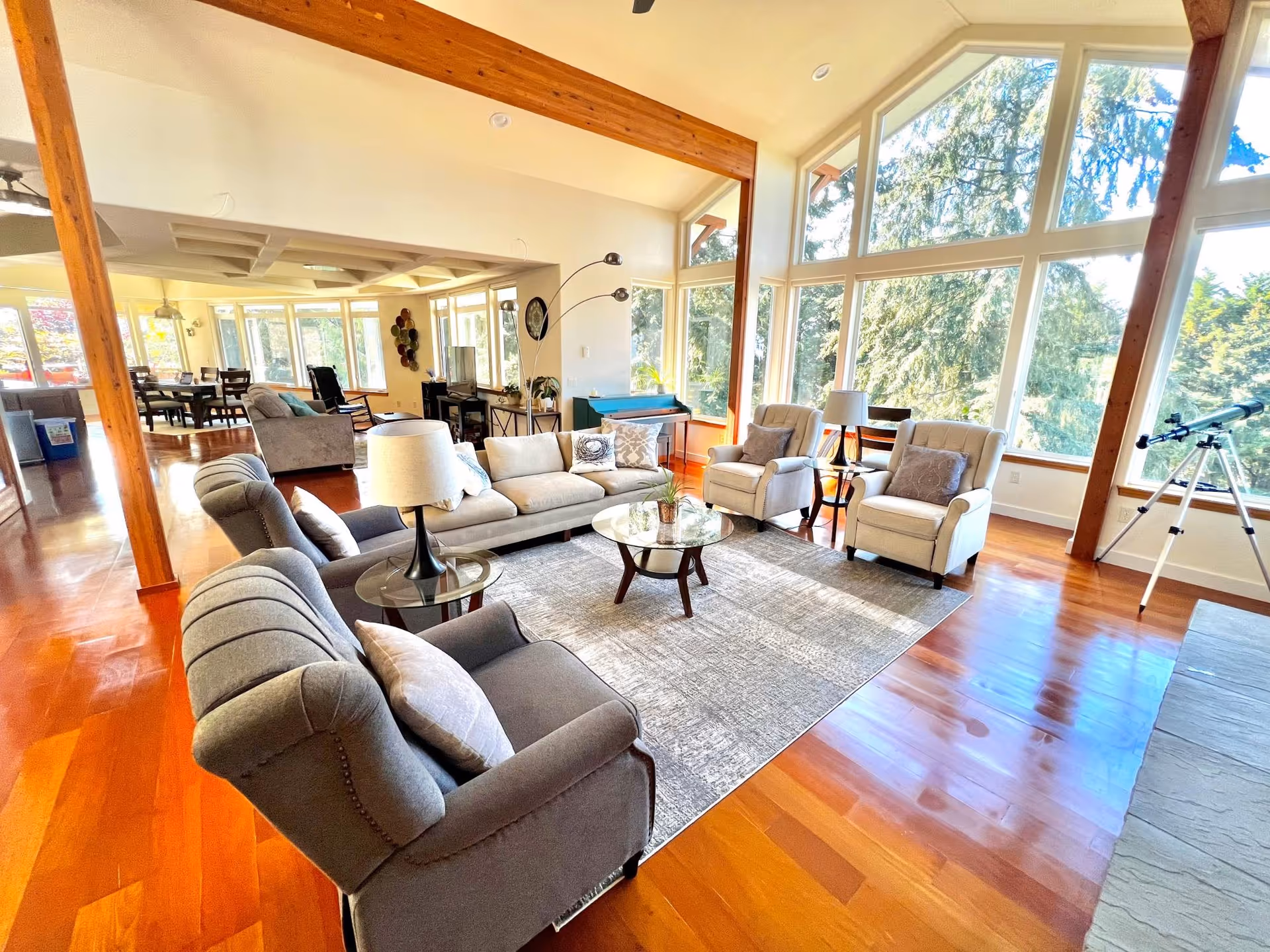 A spacious living room with large floor-to-ceiling windows letting in natural light and offering a view of trees outside. The room features wooden beams, hardwood floors, a gray couch, two beige armchairs, a gray armchair, a glass coffee table on a gray area rug, and side tables with lamps. In the background, there is a dining area with a table and chairs.