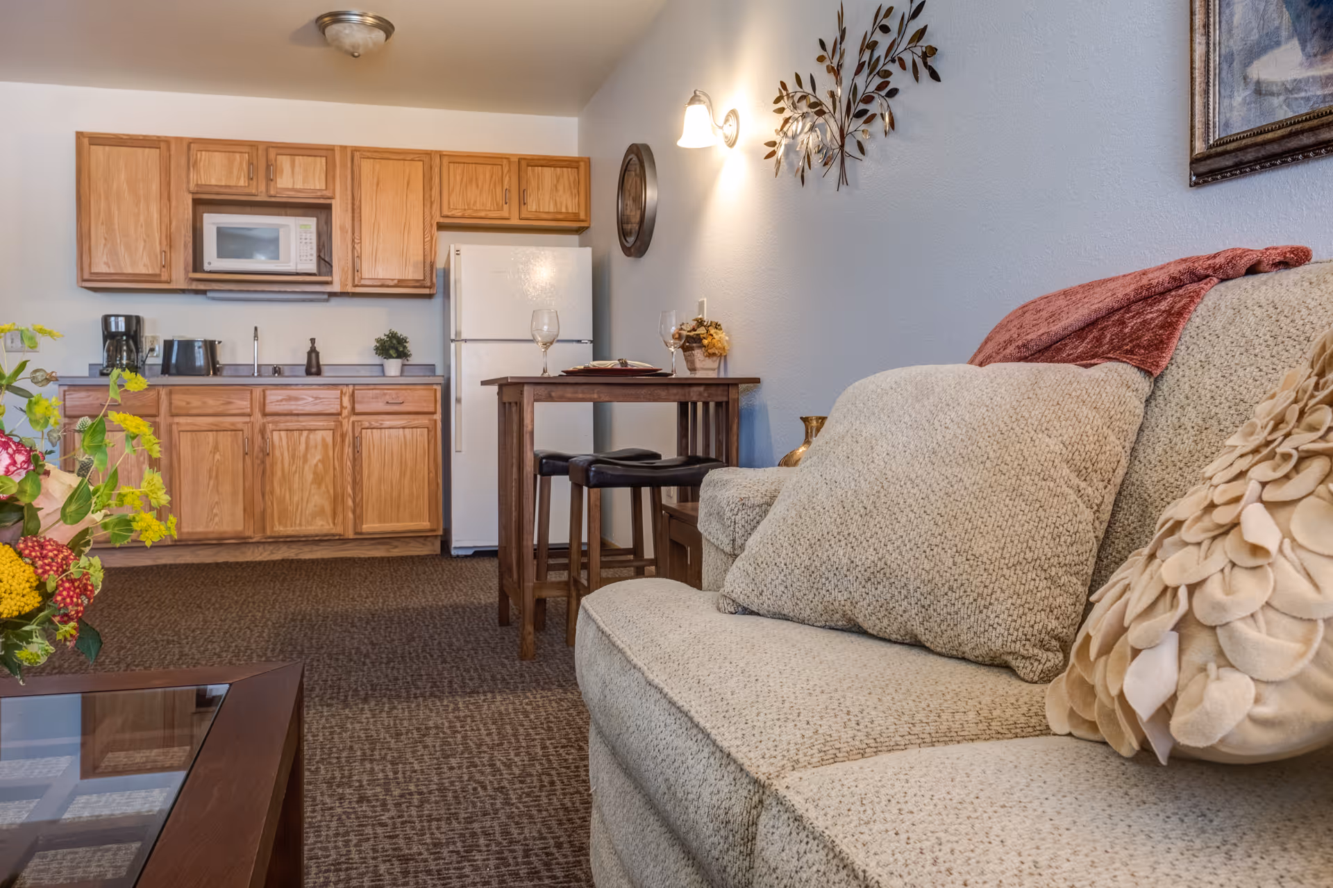 Interior view of a cozy assisted living apartment featuring a beige sofa with decorative pillows and a red throw blanket, a small wooden dining table set with two wine glasses and a plate, and a kitchenette with wooden cabinets, a white refrigerator, microwave, coffee maker, and toaster. The room is warmly lit with wall sconces and decorated with a wall clock and metal leaf art.