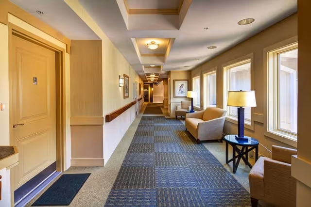 A well-lit hallway in a senior living facility with beige walls, patterned carpet, and several windows on the right side letting in natural light. The hallway features seating areas with armchairs and side tables with lamps. There are doors on the left side and framed artwork on the walls.