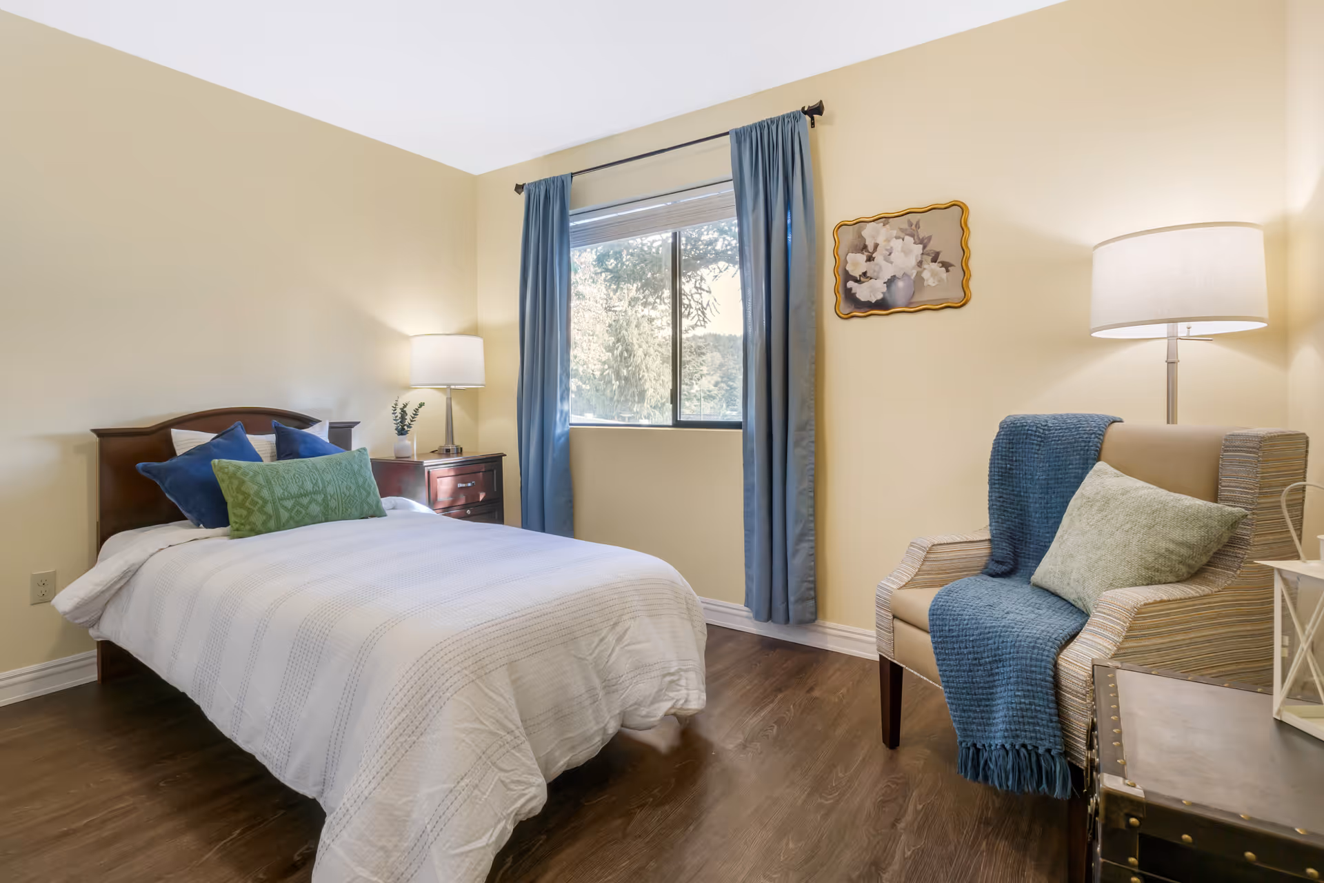 A cozy bedroom with a single bed covered in a white quilt and decorated with green and blue pillows. Next to the bed is a wooden nightstand with a lamp and a small plant. A window with blue curtains lets in natural light. Across from the bed is a comfortable armchair draped with a blue throw and a green pillow. A floor lamp and a framed floral painting adorn the wall.