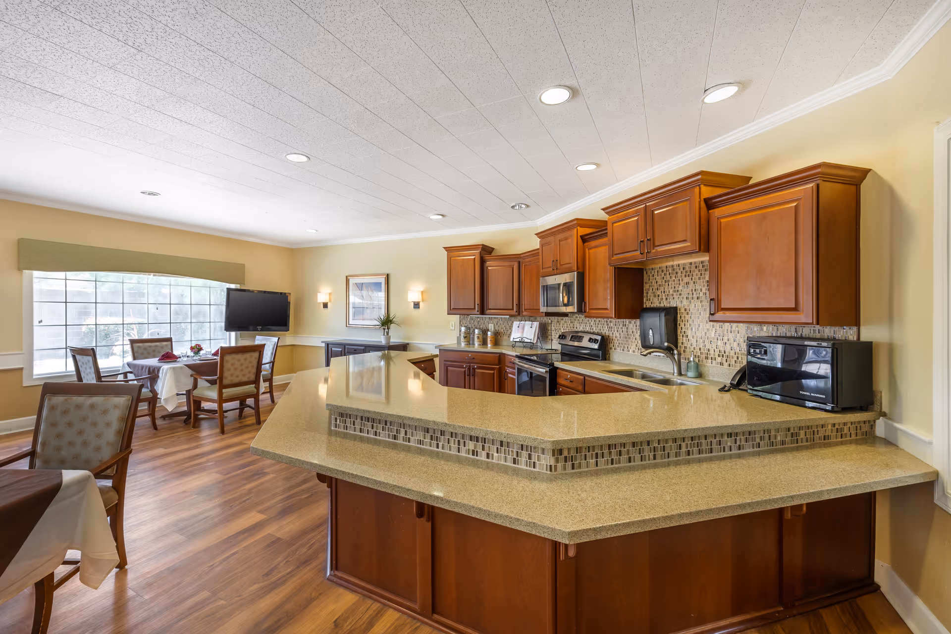 Communal kitchen and dining area with a large central island, wooden cabinets, and tables with chairs by a window.