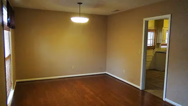Empty room with wooden floor, beige walls, a ceiling light fixture, and a doorway leading to a kitchen area with white cabinets and tiled floor.