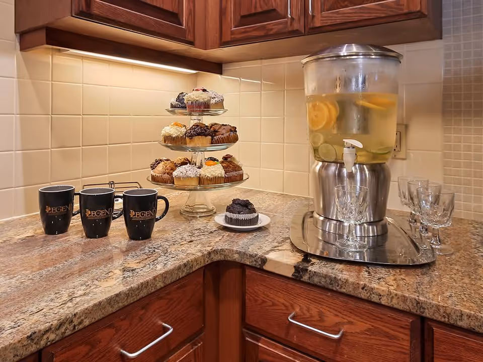 Granite countertop in a kitchen area with a three-tier tray of cupcakes, a lemon water dispenser, and three black mugs labeled 'Legend'.