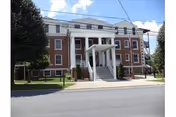 Front exterior view of a three-story brick building with white columns and a covered entrance, surrounded by a black metal fence and some trees, under a partly cloudy sky.
