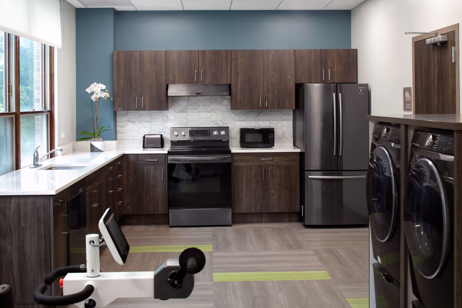 Communal kitchen featuring dark wood cabinets, a stainless steel range and fridge, white countertops by a window, and laundry machines visible on the right.