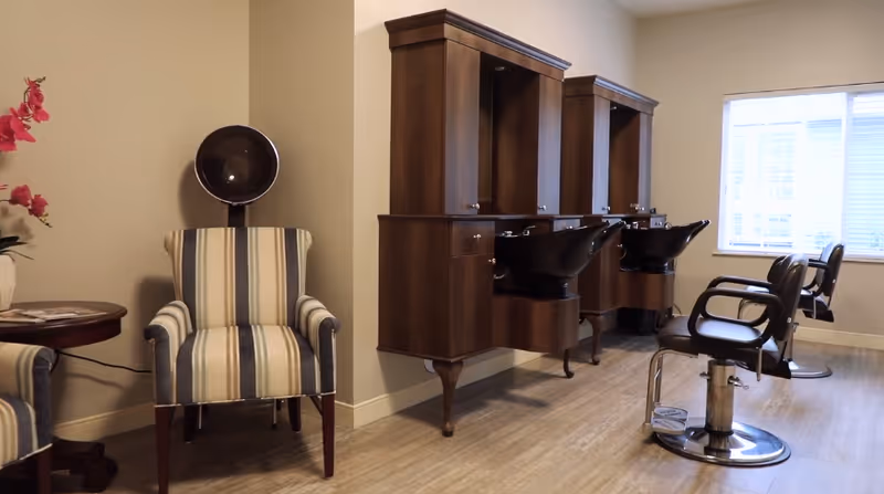 Interior view of a salon area in a senior living facility featuring two striped armchairs with a hair dryer hood between them on the left, and two black salon chairs in front of wooden wash stations with sinks on the right. A window with blinds is visible in the background.