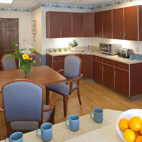 Senior living facility kitchenette with wooden cabinets, a table, chairs, and mugs on the counter.
