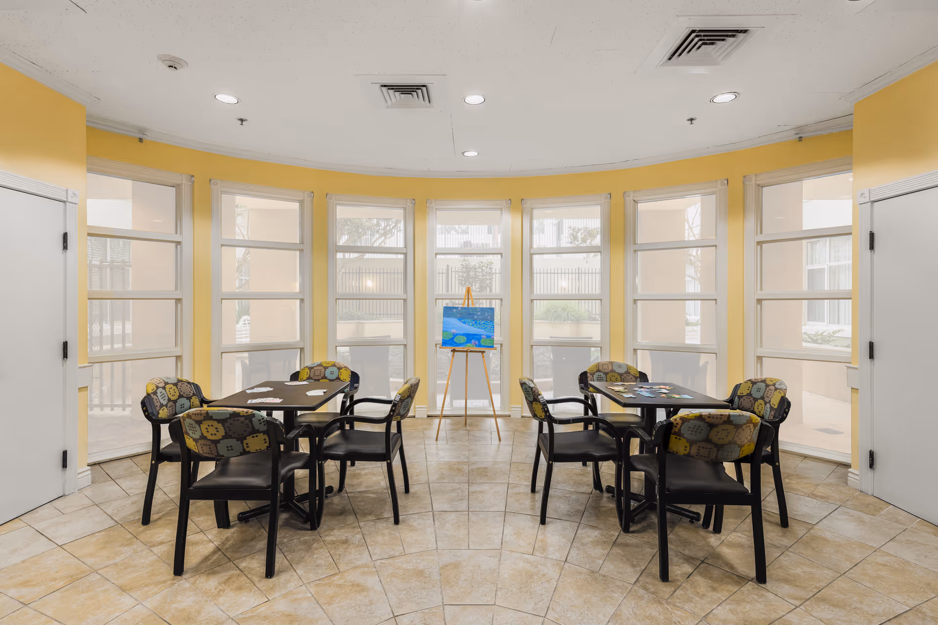 A bright room with yellow walls and large windows arranged in a curved wall. The room contains two black tables, each surrounded by four chairs with patterned cushions. On the tables are card games and puzzles. In the center near the windows, there is an easel holding a colorful painting. The floor is tiled in a beige pattern.
