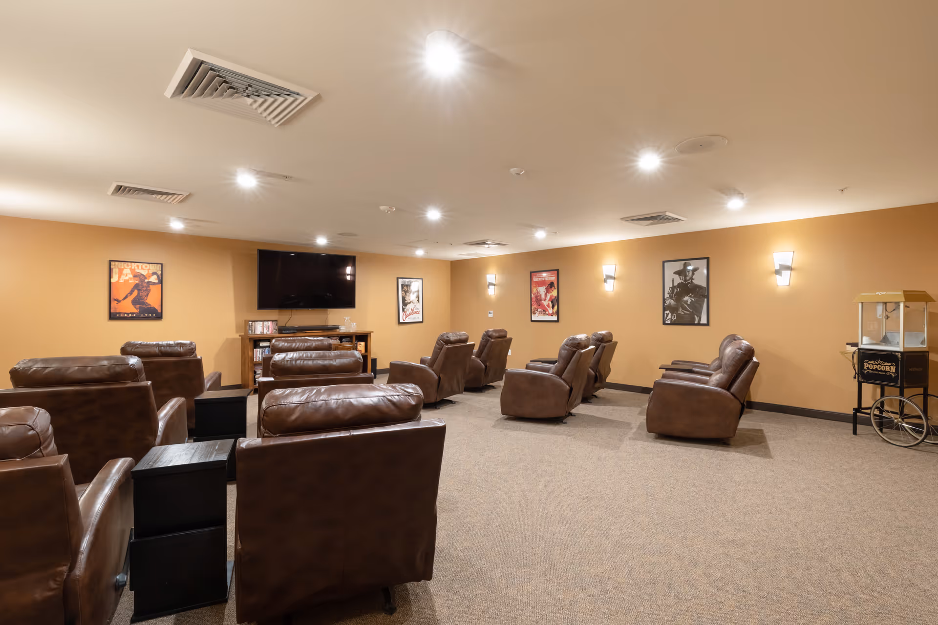 A cozy senior living facility media room with multiple brown leather recliners arranged in rows facing a wall-mounted flat-screen TV. The walls are painted a warm beige color and decorated with framed movie posters. There is a popcorn machine on a stand in the corner of the room, and the ceiling has recessed lighting.