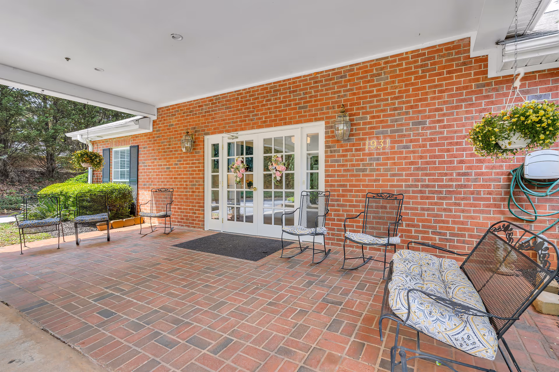 Covered outdoor patio area with brick flooring and a red brick wall featuring white double doors decorated with floral wreaths. Several metal chairs with cushions and a bench are arranged around the patio. Hanging flower pots and greenery are visible, along with outdoor wall lanterns and a garden hose mounted on the wall.