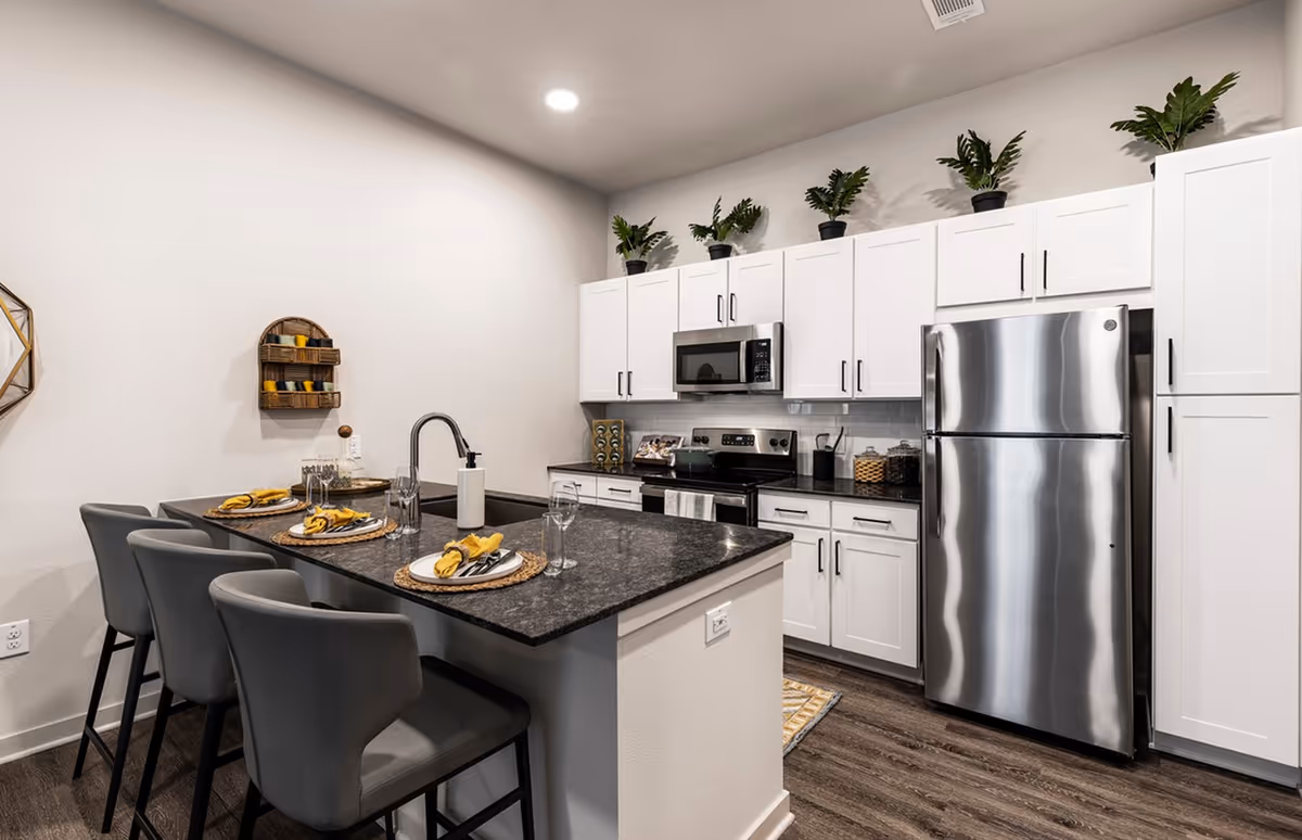 Modern kitchen with a dark island breakfast bar set for three, stainless steel appliances, white cabinets, and potted plants on top of the cabinets.