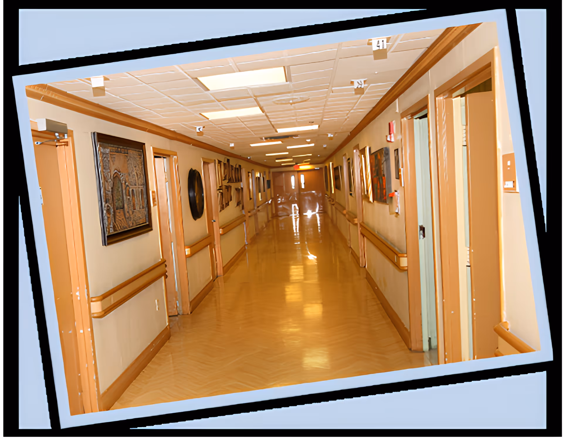 A long, well-lit hallway in a healthcare facility with beige walls, wooden handrails, and several doors on both sides. The floor is polished and shiny, and various framed artworks and decorations hang on the walls. The ceiling has recessed lighting panels.