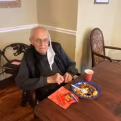 An elderly man sitting at a wooden table in a room, with a walker nearby. On the table in front of him are a plate with some food, a red cup, and a small snack pack. The man is wearing glasses and a dark sweater, and has a mask hanging from one ear.